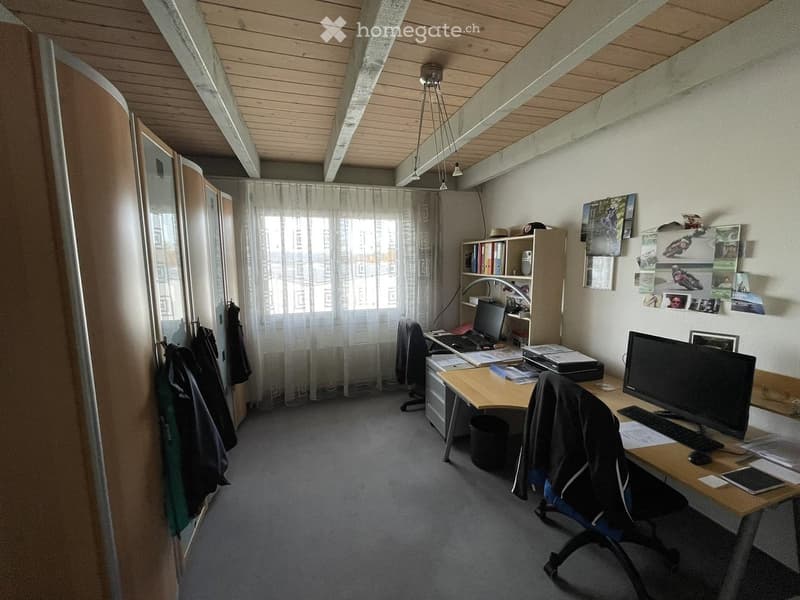 Office space with wooden ceiling, large window with white curtains, two desks with computers, chairs, shelves, and various office supplies.