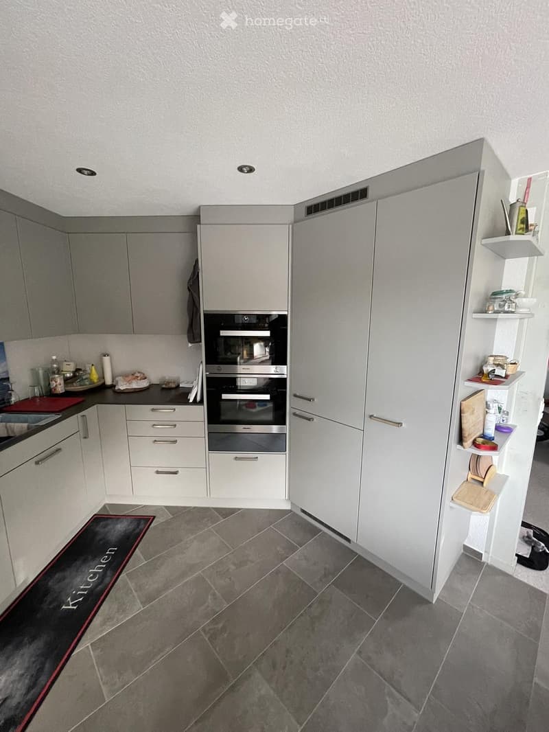 Modern kitchen with grey cabinets, stainless steel oven, white shelves, and various kitchen items on the counter and shelves.