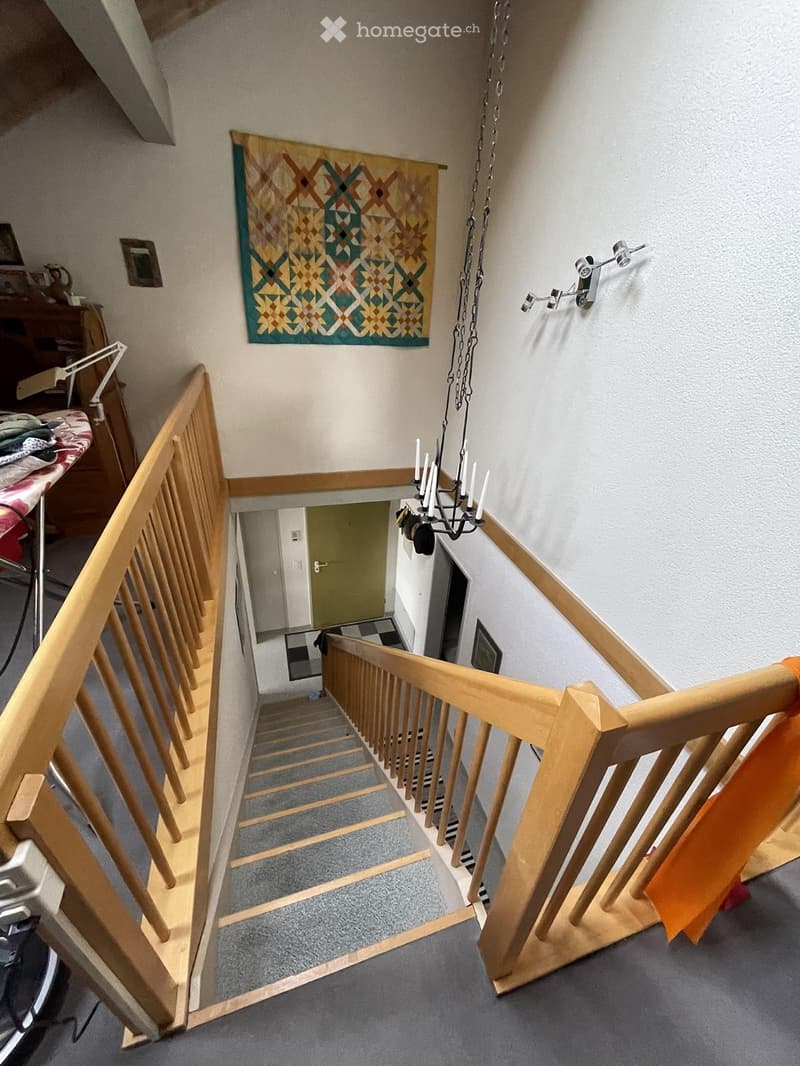 Wooden staircase with a grey carpeted step, wooden handrail, and a hanging chandelier with multiple candles. A colorful quilt hangs on the wall above the stairs, and an orange towel is draped over the railing.