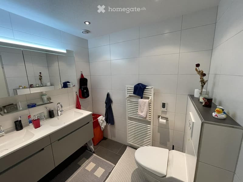 Modern bathroom with white tiled walls, a double sink, a large mirror, a toilet, a towel rack with towels, and a small decorative flower vase on the counter.