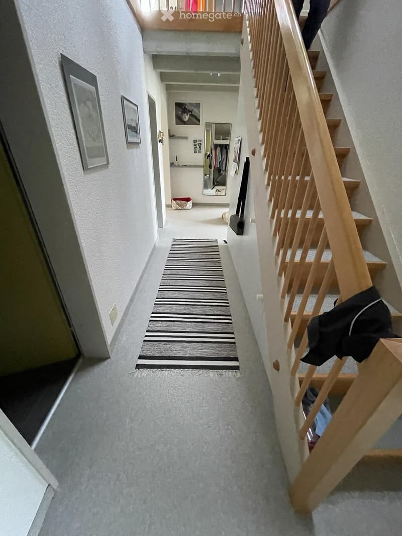 Carpeted hallway with a wooden staircase on the right, a striped runner, framed pictures on the walls, and an open closet with clothes and a pet bed visible at the end.