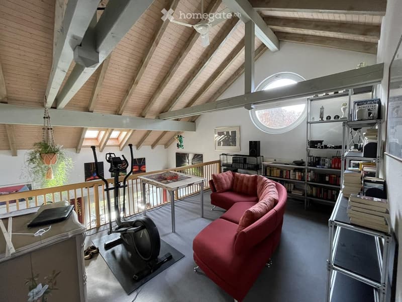 Loft-style room with exposed wooden beams, skylights, a red sectional sofa, a small table, a bookshelf, an exercise bike, and a hanging plant.