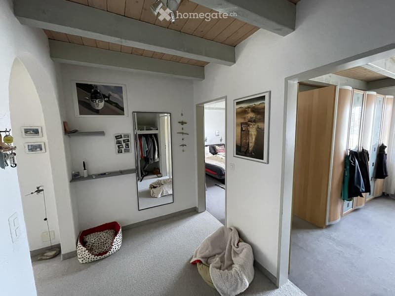 Modern, minimalist room with white walls, carpeted floor, large mirror, bed visible through doorway, picture frames, dog bed, and a beige chair.