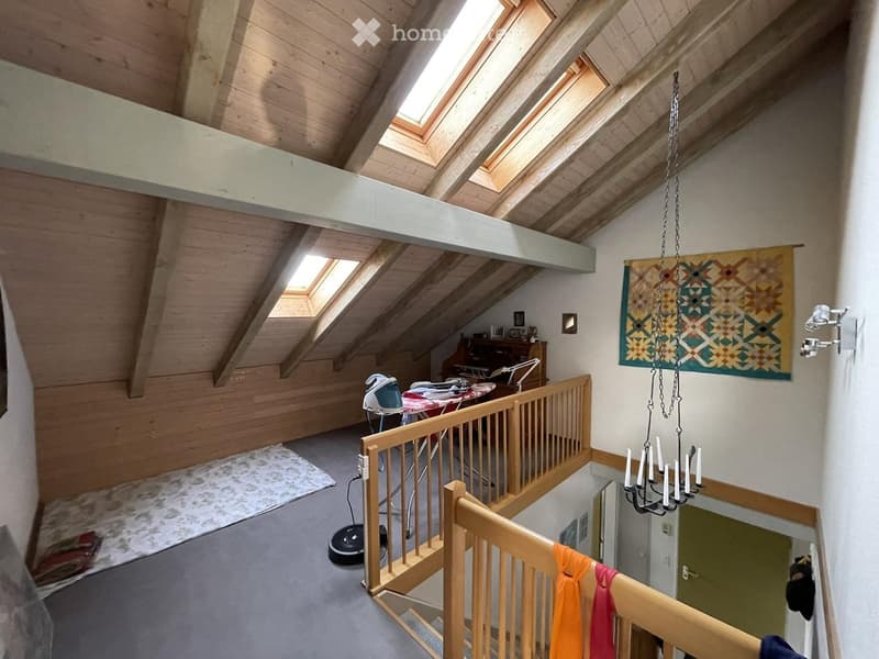 Loft-style room with wooden beams, skylights, and a railing overlooking a lower level. Contains an ironing board, iron, table, chair, lamp, mirror, and a decorative chandelier with a colorful tapestry.