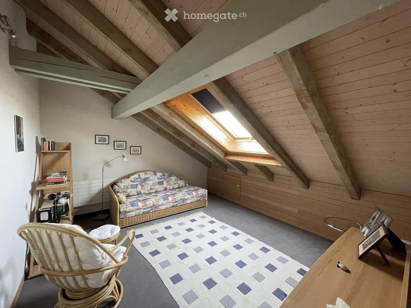 Cozy attic living space with exposed wooden beams, skylights, a patterned sofa, wicker chair, coffee table, bookshelf, and floor lamp.