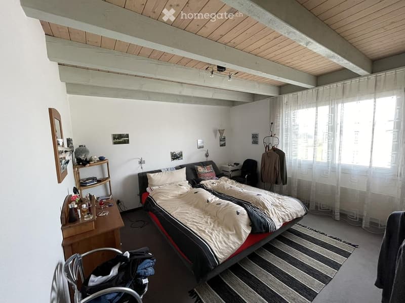 Cozy bedroom with a double bed, nightstand, chair, mirror, and shelf. The room features exposed wooden beams on the ceiling and large windows with sheer curtains.