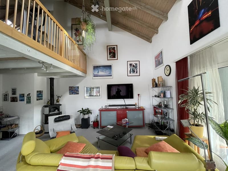 Bright living room with a green sofa, coffee table, television, fireplace, and decorative plants. The room features exposed wooden beams and a staircase leading to an upper level.