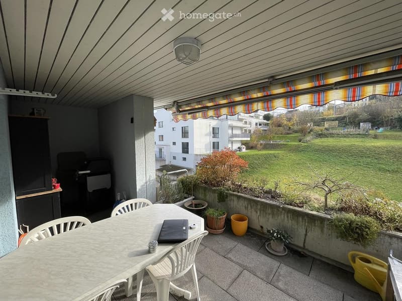Balcony with a table, chairs, laptop, potted plants, and a view of a green field and buildings.