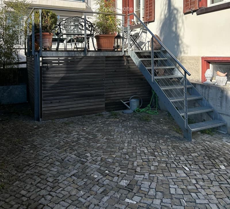 Garden area with cobblestone ground, metal staircase leading to a raised terrace, potted plants, and outdoor furniture.
