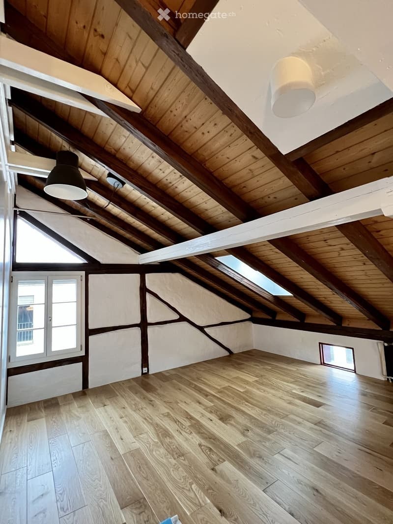 An empty attic space with wooden beams, exposed wooden ceiling, wooden floor, and skylights. The walls are white with a textured finish, and there are two windows on the left side.