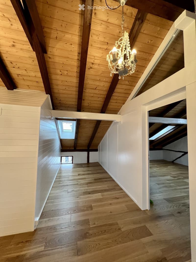 Bright, airy hallway with wooden flooring and exposed wooden beams on the ceiling. A chandelier hangs from the ceiling, and there are skylights providing natural light. The walls are painted white, and the space is open and uncluttered.