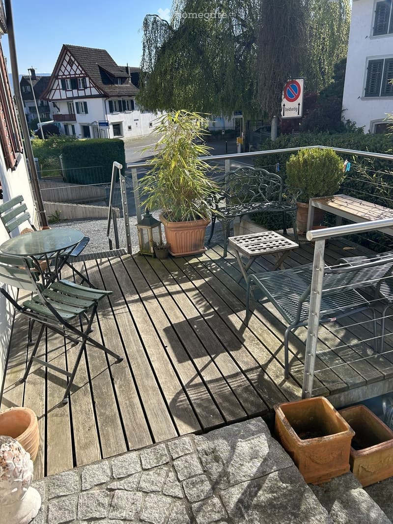 Balcony with wooden decking, metal railing, outdoor furniture including a table, chairs, bench, lantern, and potted plants.
