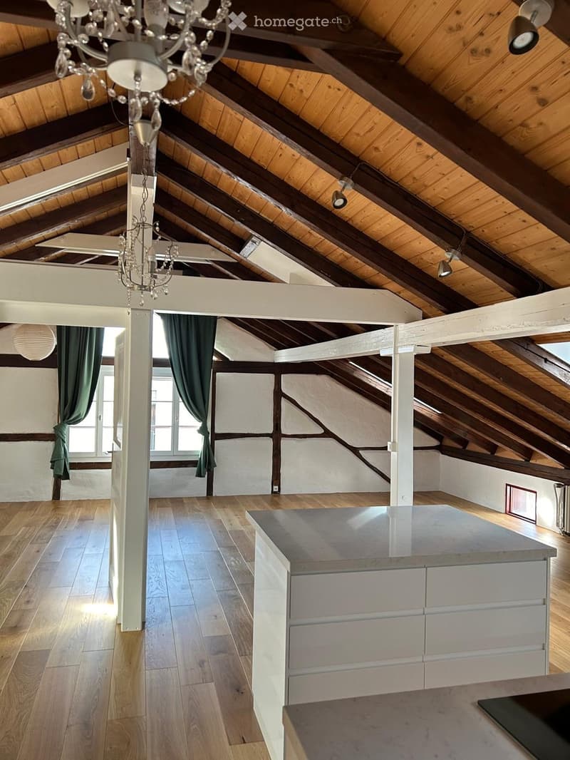 Open-plan attic space with exposed wooden beams, wooden flooring, and a modern kitchen island. Green curtains on windows, and a chandelier hanging from the ceiling.