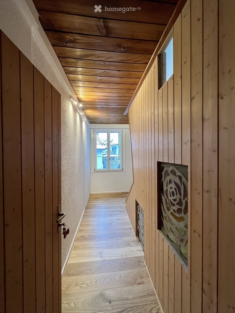 Wooden-paneled hallway with a slanted wooden ceiling, recessed lighting, and a window at the end. Features a decorative stained-glass panel on one wall and a door with a keyhole on the left.