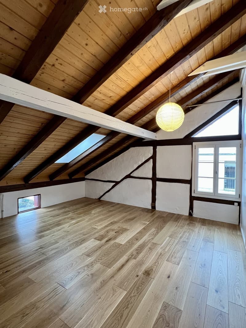 Empty attic space with wooden floors, exposed wooden beams, skylights, and large windows, featuring white walls and a hanging light fixture.