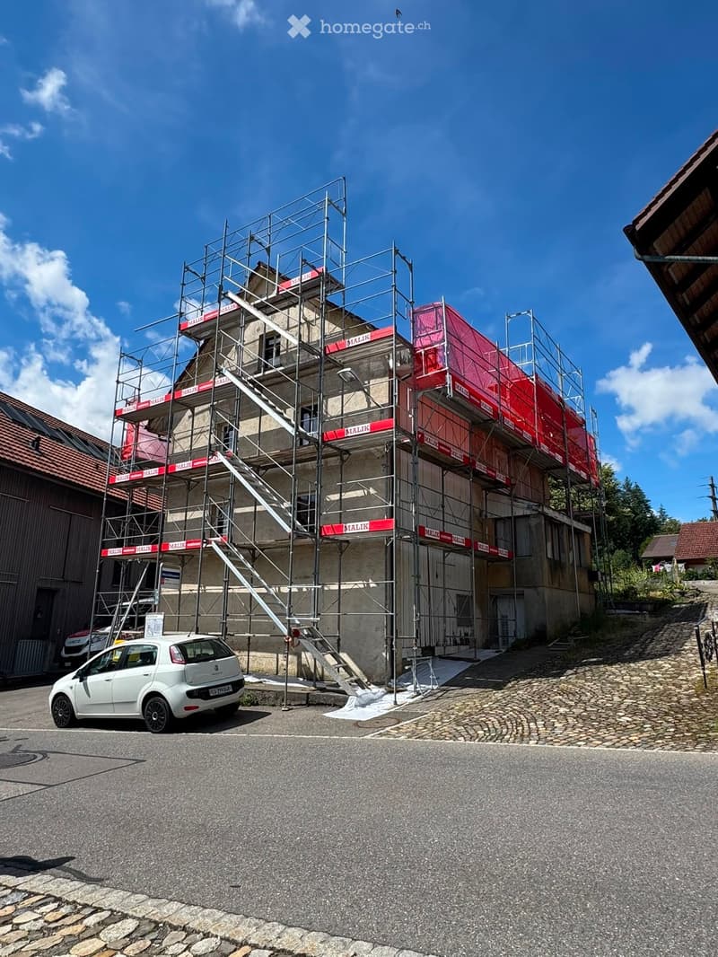 A multi-story building under construction with scaffolding, red protective covers, and a staircase leading to the upper levels. A white car is parked in front.