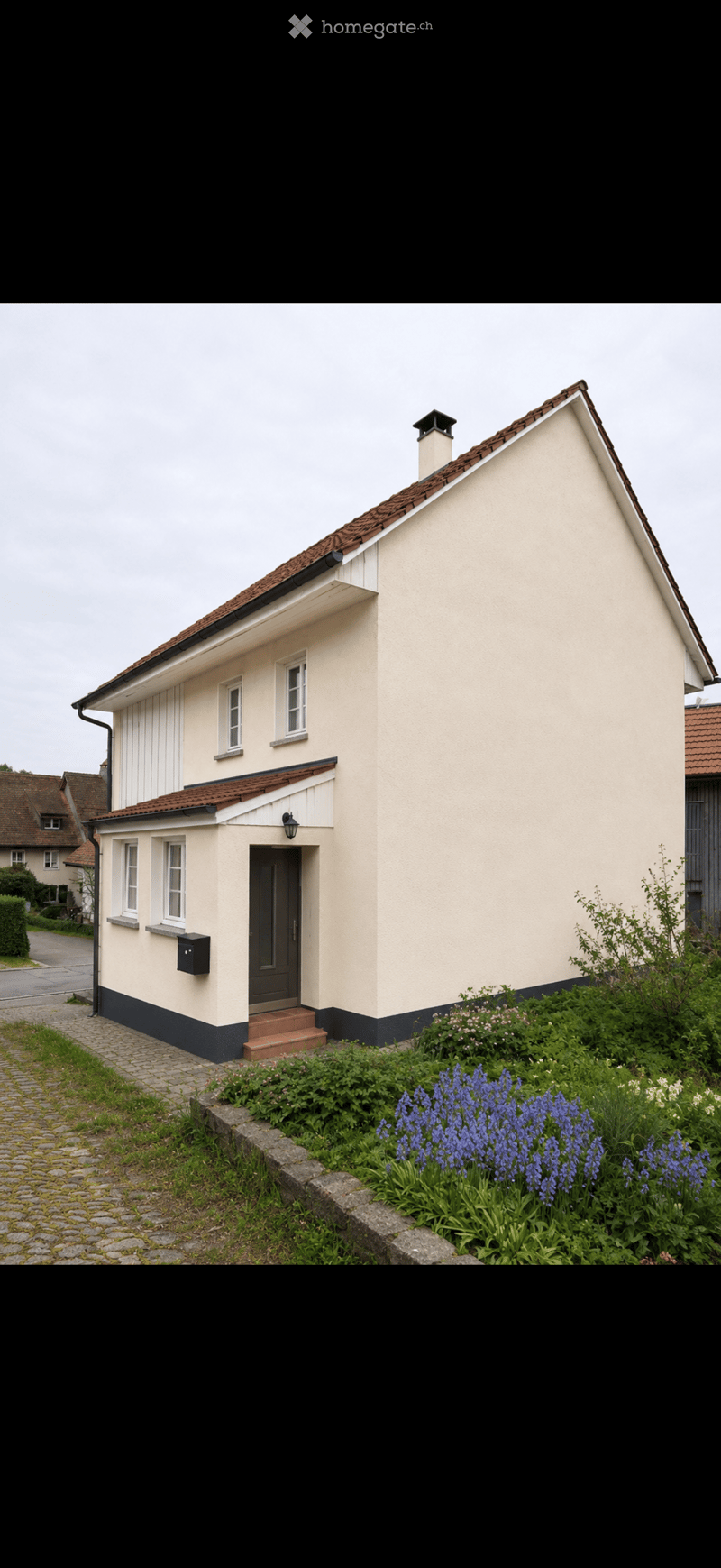 Single-story house with beige walls, dark blue trim, red-tiled roof, and a small chimney. Features a front door with a small step, windows, and a garden with blue flowers and shrubs.
