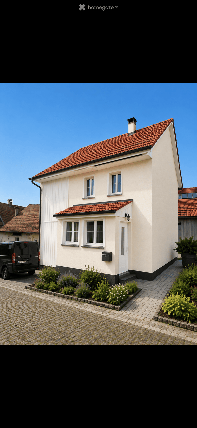 Single-story house with white walls, red-tiled roof, multiple windows, and a small garden area with plants and shrubs.