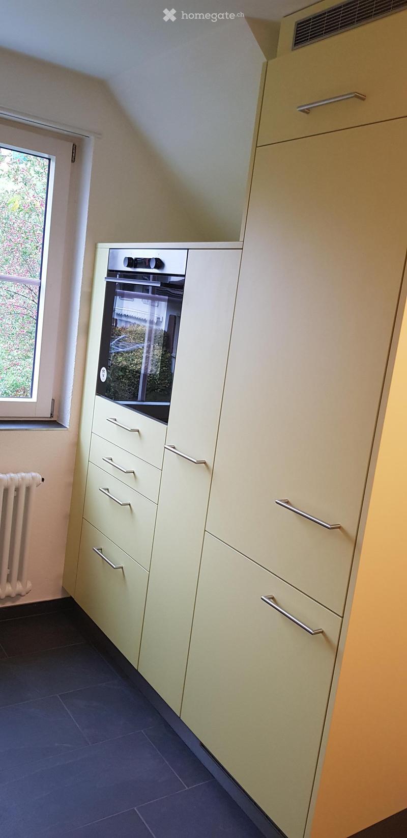Modern kitchen with light-colored cabinets, stainless steel handles, and a built-in oven. Dark tiled floor and a radiator visible on the left side.