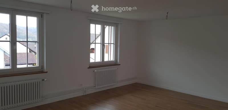 Empty room with wooden flooring, white walls, and two windows offering a view of neighboring houses. Radiators are present under each window.