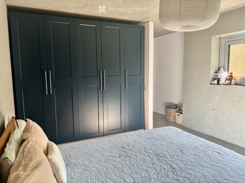 Modern bedroom with a large dark green wardrobe, a bed with light-colored bedding and pillows, and a wicker basket on the floor near the window.