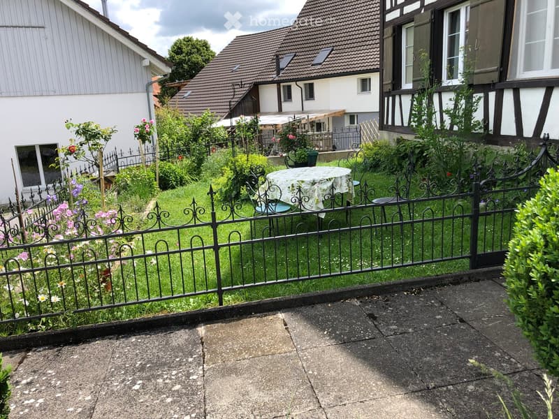 a garden with a fence, table, chairs, green grass, flowers, plants, bushes, trees