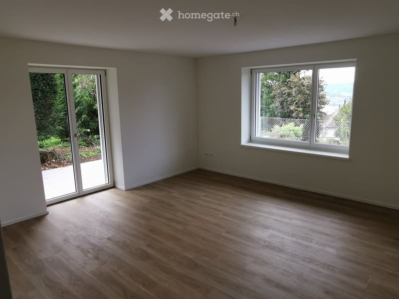 Empty room with light wooden flooring, white walls, large windows, and a sliding glass door leading to an outdoor area.