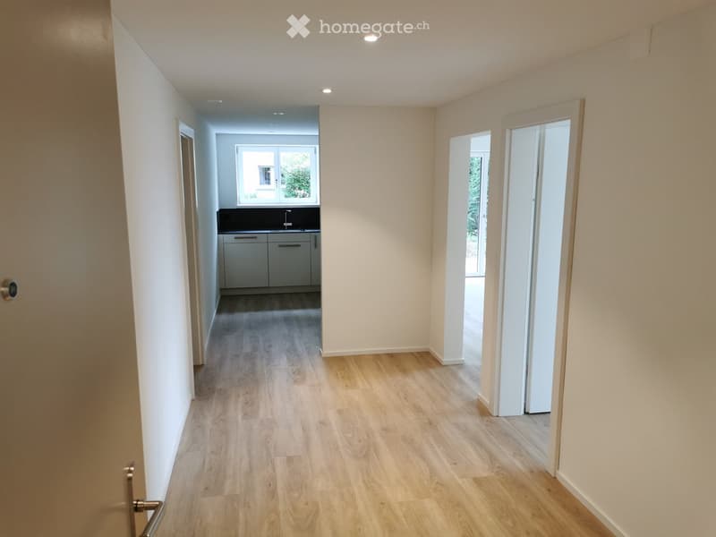 Bright hallway with light wooden flooring, white walls, and recessed lighting. Open doorways lead to a kitchen and another room.