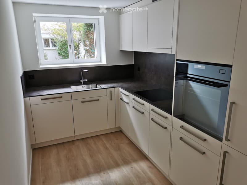 Modern kitchen with white cabinets, dark countertops, a sink, and an oven. The space features wooden flooring and a window offering a view of greenery.