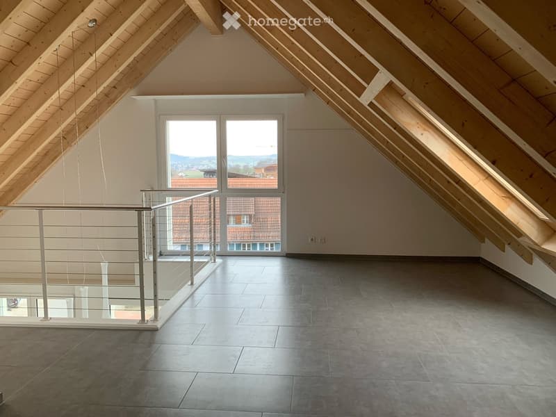 Empty attic space with wooden slanted ceilings, large windows, and a modern glass railing leading to a balcony.