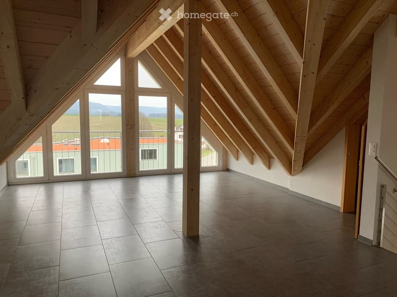 Bright, spacious attic room with wooden beams, large windows, and a balcony overlooking a green landscape.