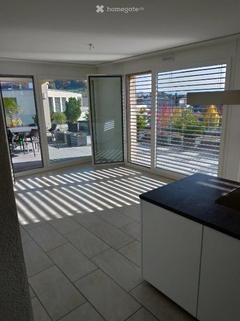 Bright, modern room with large windows and a view, featuring a tiled floor, a small kitchen area with cabinets, and a table and chairs in the adjacent outdoor space.
