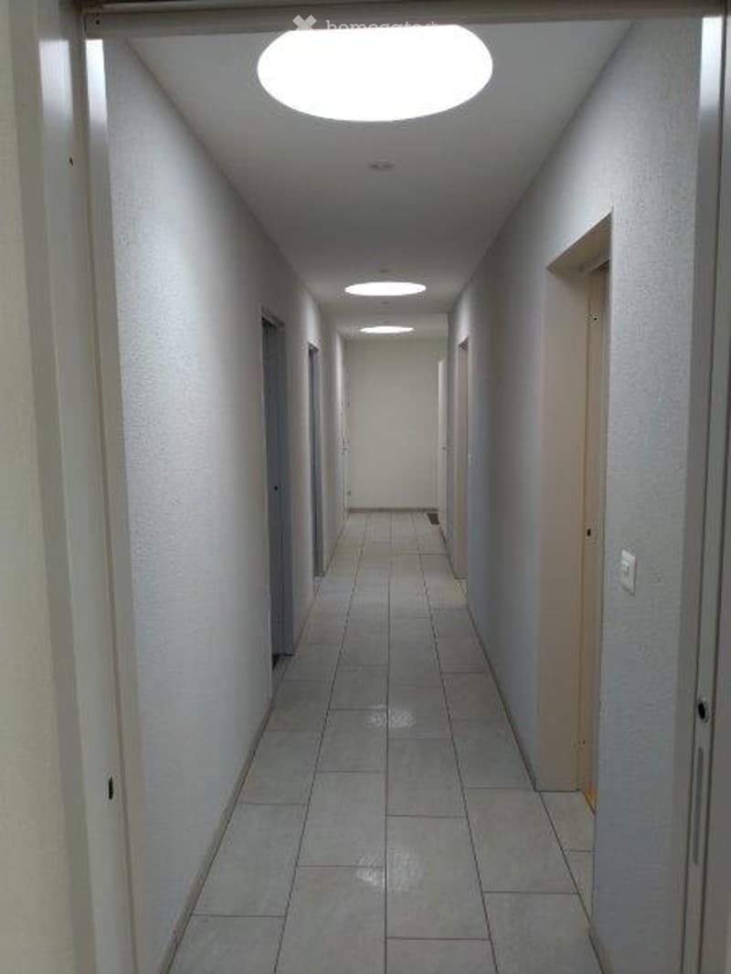 A clean, well-lit hallway with tiled flooring and white walls, featuring several doors on both sides and recessed ceiling lights.