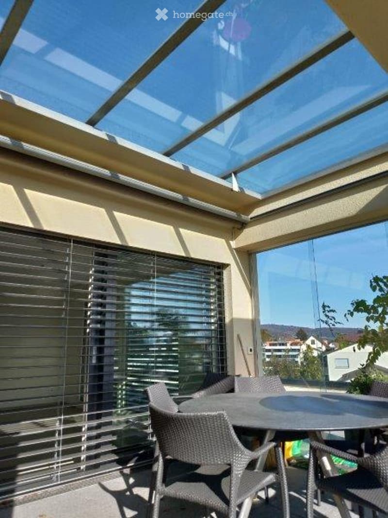 A modern, enclosed terrace with a glass roof and large windows, featuring a round table and several wicker chairs.