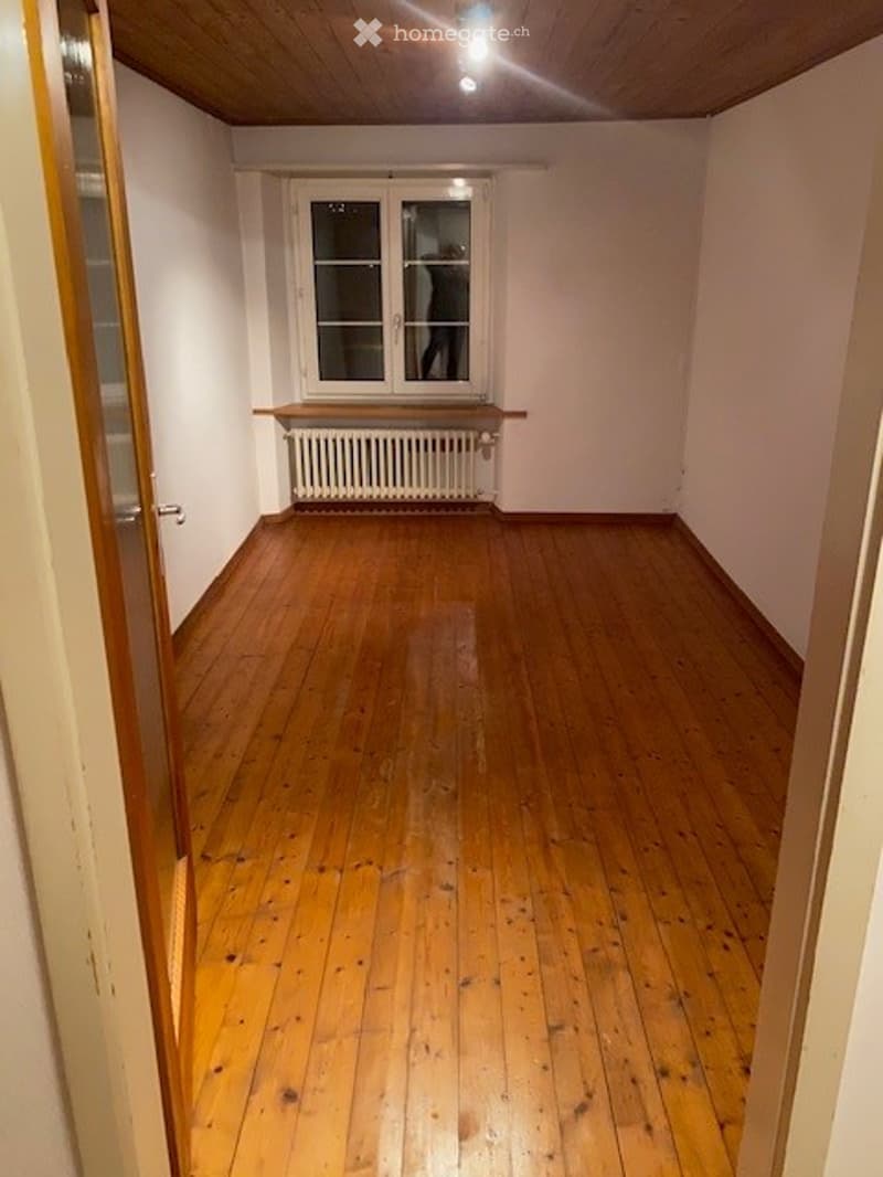 Empty room with wooden flooring, white walls, and a window with a radiator below it. The room has a simple, clean design with no furniture or decorations.