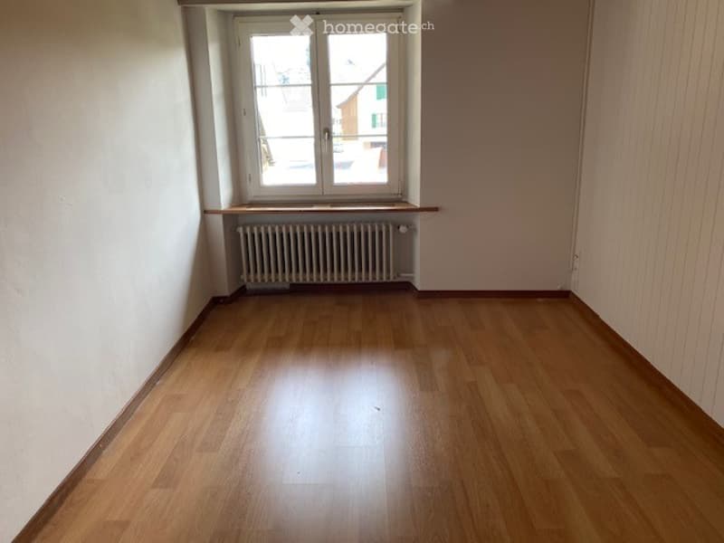 Empty room with wooden flooring, white walls, a window with white frame, and a radiator below the window.