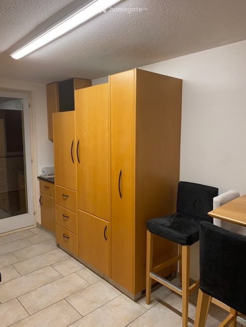 A small, furnished room with a large wooden cabinet, a black stool, a wooden chair, and a small table.