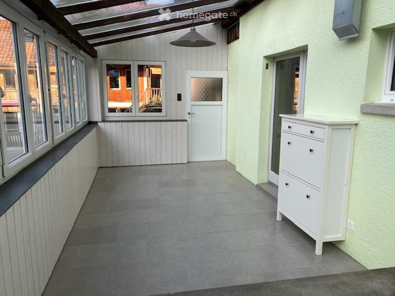 Bright, enclosed patio area with grey tiled flooring, white wooden paneling, large windows, and a white dresser with drawers.