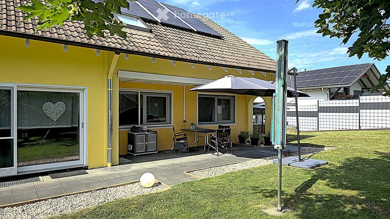 Bright yellow house with a tiled roof, solar panels, and a covered patio area with a table, chairs, and an umbrella. The garden features a well-maintained lawn, a small gravel area, and a decorative ball.