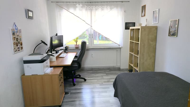 A well-lit room with a desk setup, a comfortable chair, a printer, and shelves. The room features a bed with a dark blanket, a window with white curtains, and several picture frames on the walls.