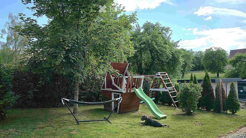 A well-maintained garden with a wooden playhouse, green slide, swing set, hammock, and a dog resting on the grass.