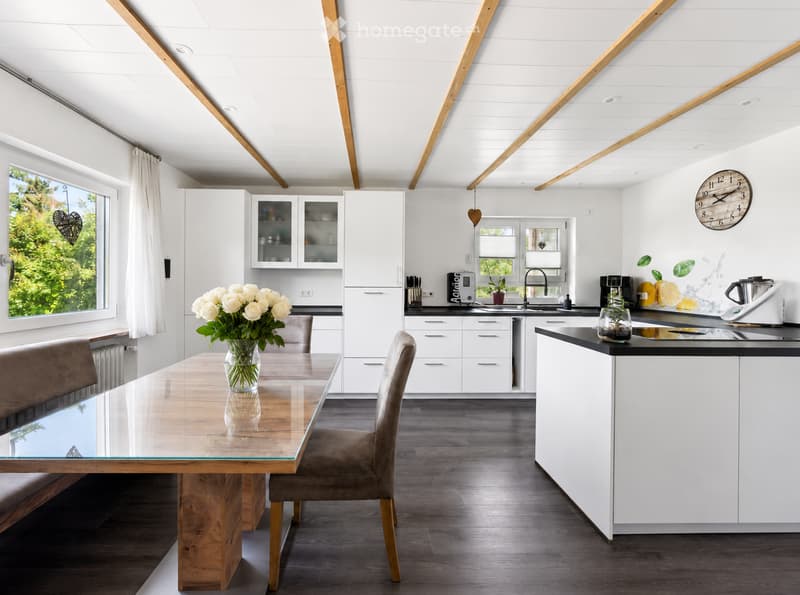 Modern kitchen with white cabinets, dark countertops, a wooden dining table with glass top, two chairs, and a bench. Features include a sink, faucet, coffee maker, blender, and a decorative vase with white roses on the table.