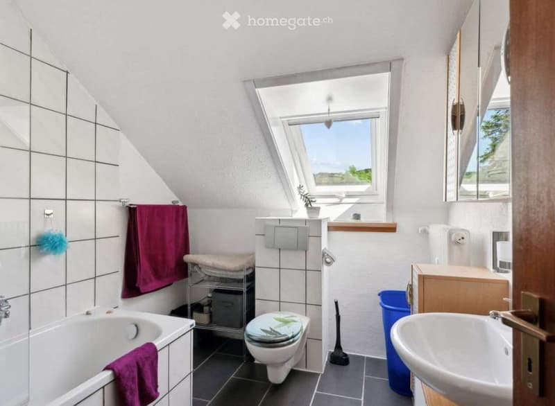 Bright bathroom with a slanted ceiling, large window, bathtub, toilet, sink, mirror, towel rack, and storage shelf.