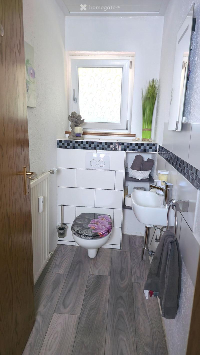 Modern bathroom with wooden flooring, white tiles, a toilet with a floral seat cover, a sink with a chrome faucet, and a small shelf with towels and toiletries.