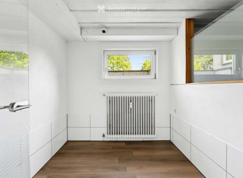 Completely empty room with white walls, wooden floor, radiator, and large windows with a view of greenery.