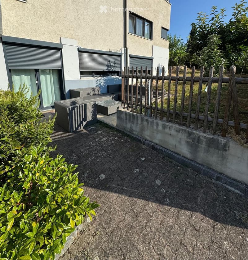 Outdoor patio area with a sofa, table, and wooden fence, adjacent to a modern building with large windows and roller shutters.