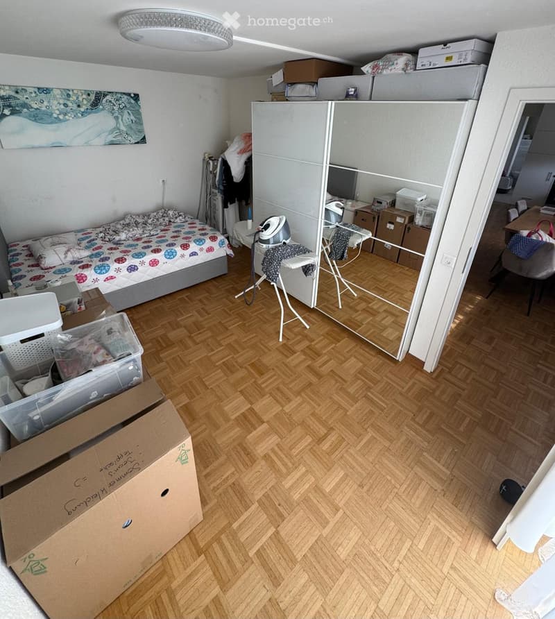 A furnished bedroom with a bed, a mirror, a refrigerator, a screen, a chair, a table, and an ironing board.