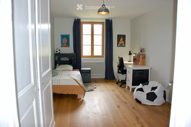 Well-lit bedroom with wooden flooring, a bed with orange and white bedding, a desk with a chair, a beanie bag, a bookshelf, and artwork on the walls.