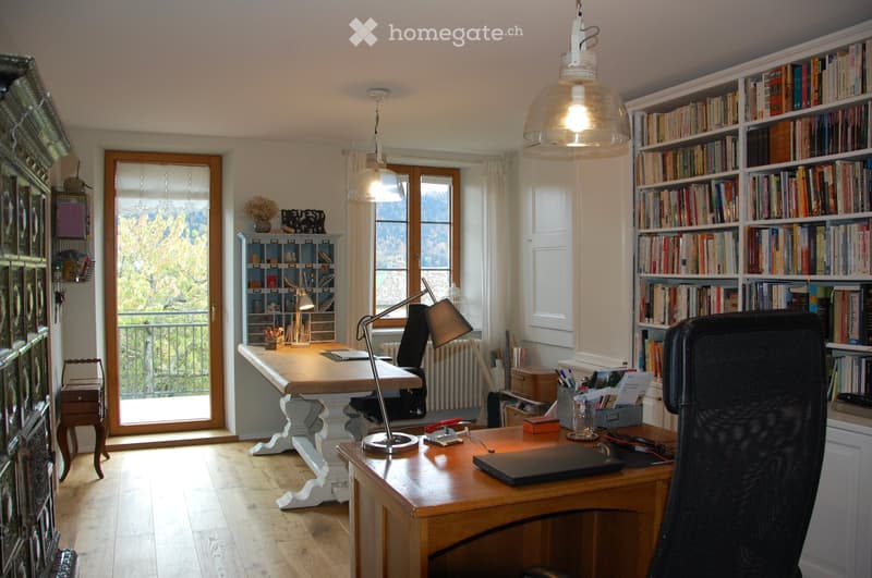 Bright, well-furnished study room with wooden flooring, large windows offering a scenic view, a large wooden desk with a laptop, a comfortable office chair, a modern lamp, and a tall bookshelf filled with books.