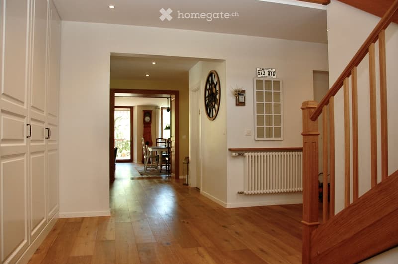 Spacious hallway with wooden flooring, white walls, and a staircase leading upwards. Features a radiator, a wall-mounted clock, and a framed address sign. A dining area is visible in the background with a table and chairs.
